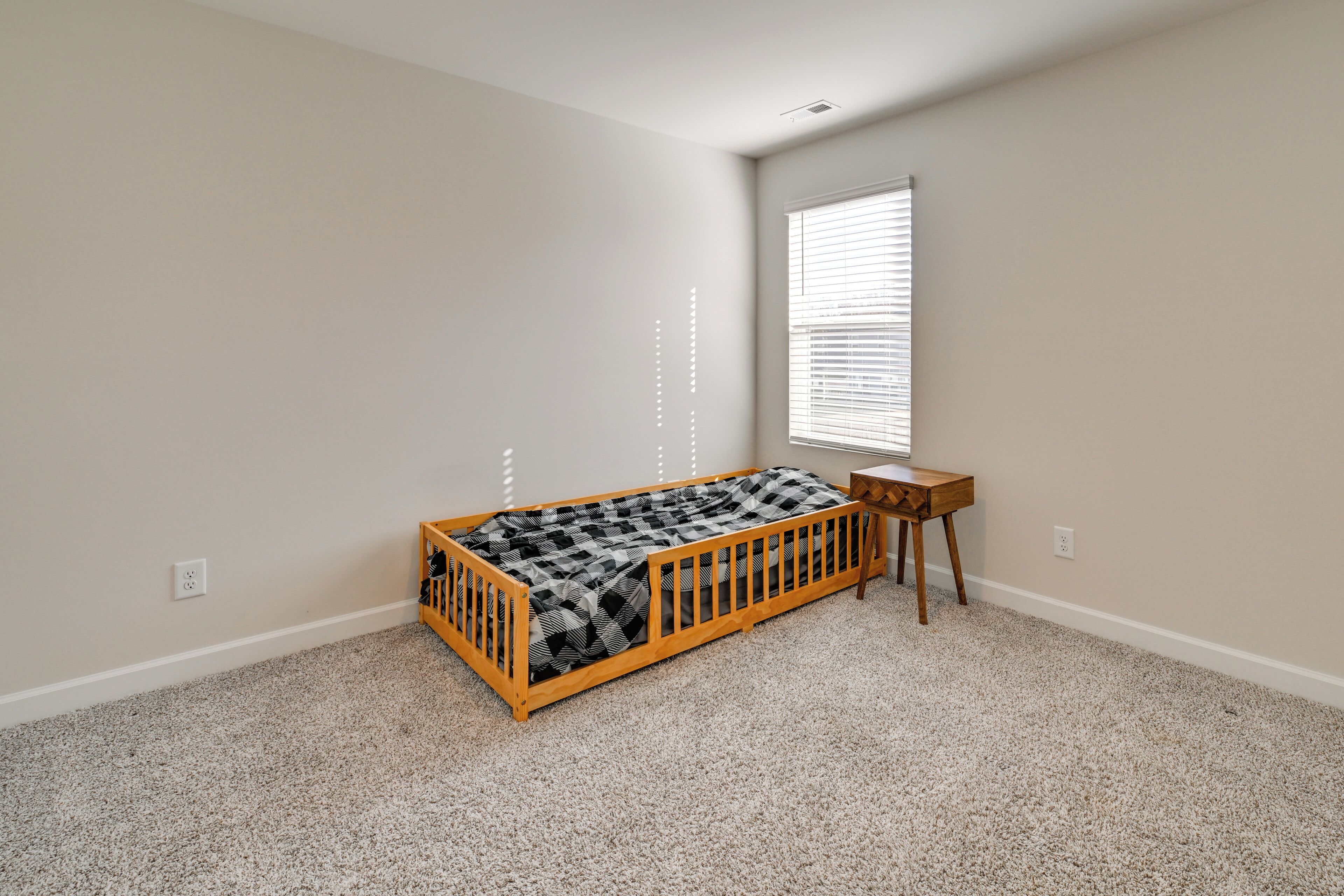 A single room with a wooden bunk bed, a small wooden nightstand, a window with blinds, and a carpeted floor.