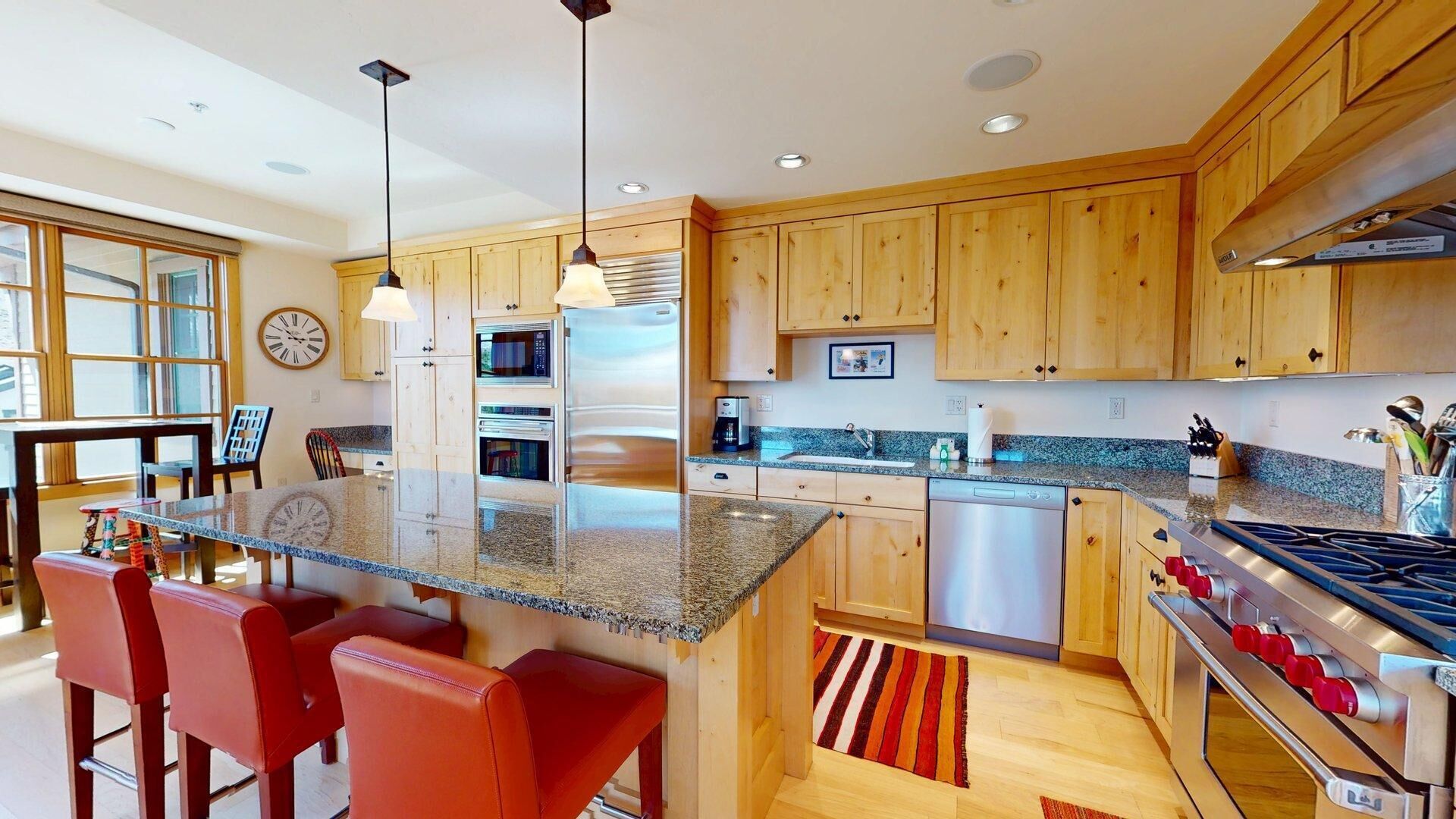 A modern kitchen with wooden cabinets, a granite countertop, stainless steel appliances, and a dining area with red chairs.