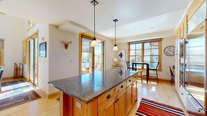 A modern kitchen with granite countertops, wooden cabinets, a central island, pendant lighting, a dining area with a table and chairs, and a large window with a view. - Elkhorn Springs 15 - Luxury w/ Great Views, Walk to Pool, Tennis, Golf (Elkhorn)