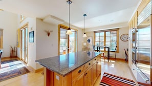 A modern kitchen with granite countertops, wooden cabinets, a central island, pendant lighting, a dining area with a table and chairs, and a large window with a view. - Elkhorn Springs 15 - Luxury w/ Great Views, Walk to Pool, Tennis, Golf (Elkhorn)