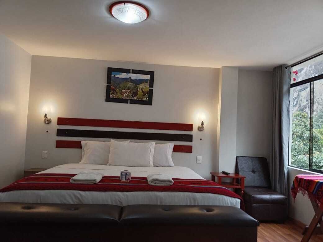 Ein Hotelzimmer mit einem großen Bett, einem roten und schwarzen Streifen-Kopfende, einem Nachttisch, einem Sessel, einem Tisch mit Tischdecke und einem Fenster mit Blick auf Grünflächen.