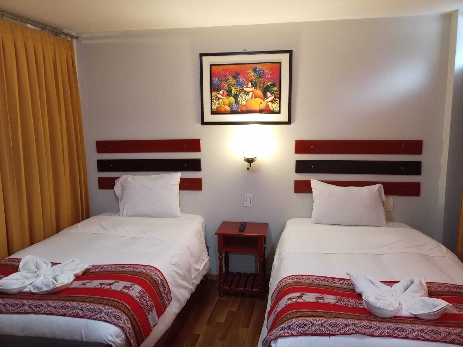 A hotel room with two beds, each with a red and white patterned bedspread, white pillows, and a small bedside table with a remote control. A framed painting hangs above the beds.