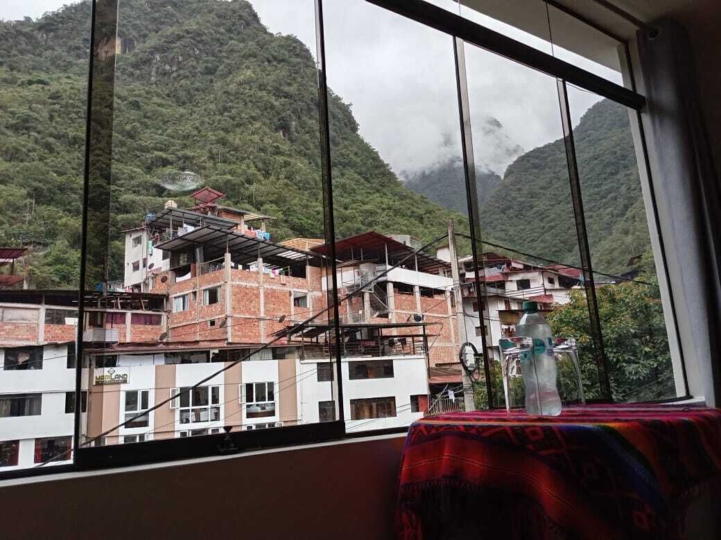 A view through a window of a building with a mountain backdrop, featuring a table with a patterned tablecloth and a bottle.