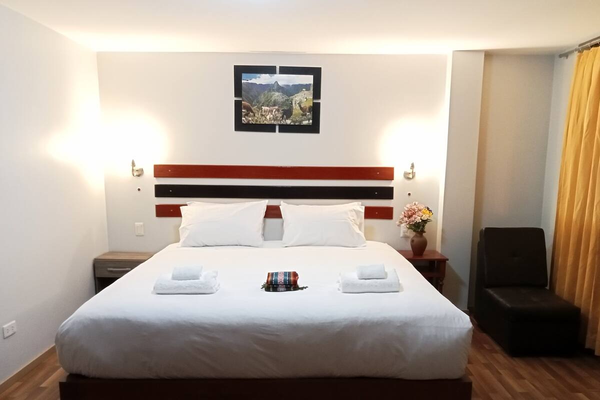A neatly made bed with white linens and two towels, a wooden headboard, a nightstand with a vase of flowers, and a framed picture on the wall.