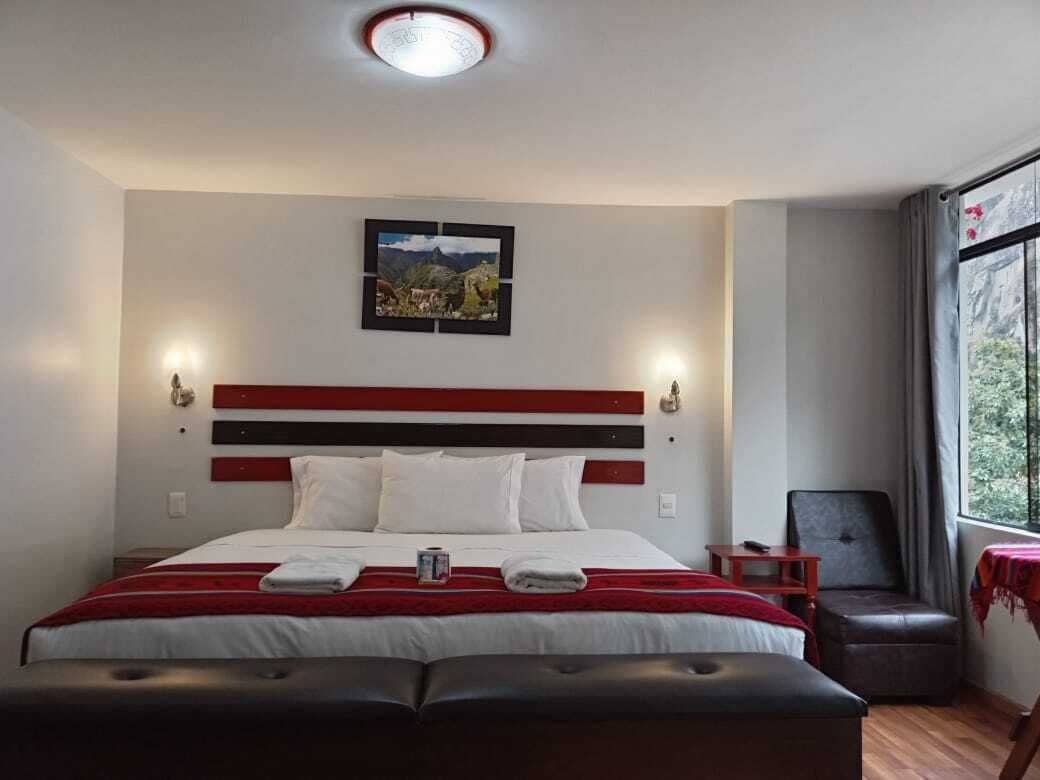 A hotel room with a large bed, a headboard with horizontal stripes, a bedside table, a chair, a window with a view of greenery, and a picture on the wall.