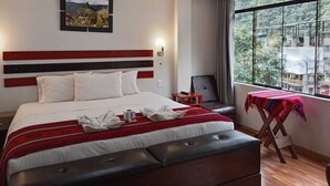 A hotel room with a large bed, a headboard with horizontal stripes, a window with a view of greenery, and a small table with a chair. - Raymi MachuPicchu (Aguas Calientes)