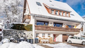 A two-story building with a snow-covered roof, a balcony with a wooden railing, and a car parked in front. - Apartment 3 with Shared Garden and Wi-Fi (Lenzkirch)