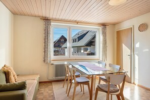 Ein Zimmer mit Holzbalkendecke, einem fenster mit Vorhängen, einem Tisch mit Stühlen, einem Heizkörper und einer Tür.