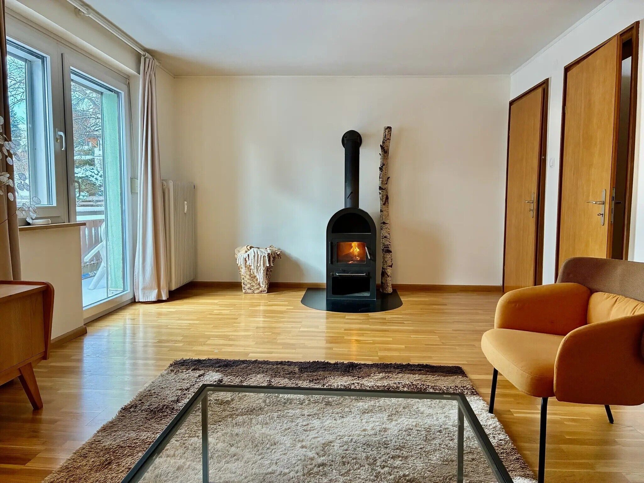Ein Wohnzimmer mit Kaminofen, Holzboden, einem Glastisch und einem modernen orangefarbenen Sessel.