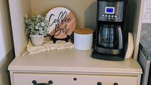 A kitchenette with a coffee maker, a sign reading 'Coffee Bar,' and stacked plates and cups on a shelf. - Hometown Haven
quiet•peaceful•cozy (Hancock)