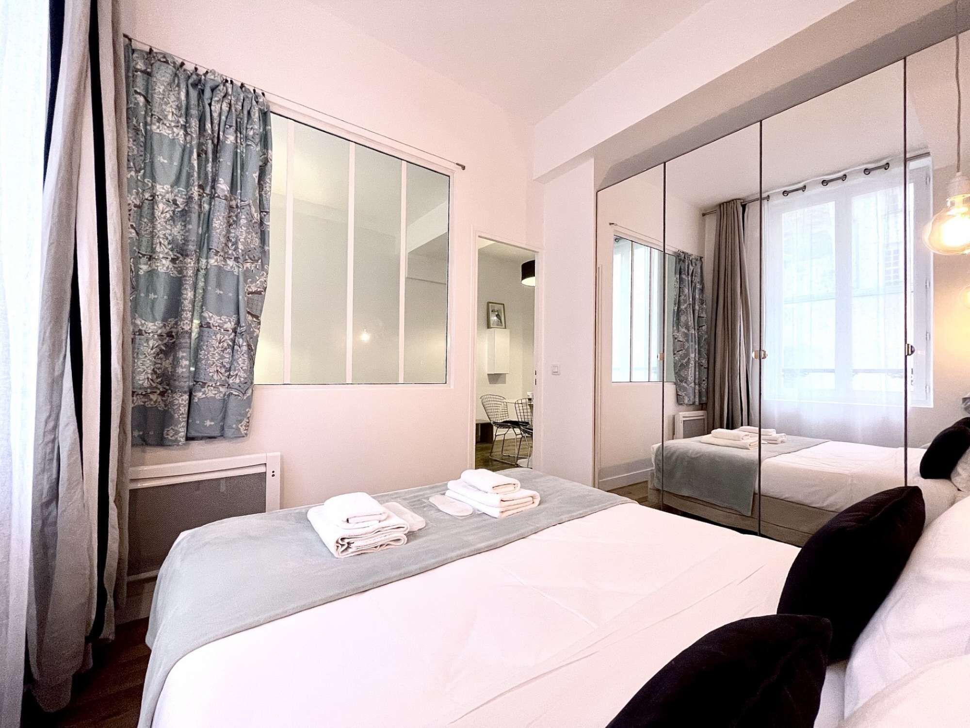 Une chambre d’hôtel avec un grand lit, un miroir, une fenêtre avec des rideaux et un petit bureau avec une chaise.