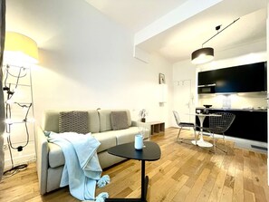 A modern living room with a sofa, a small round table, a dining area with a chair, and a kitchen area with cabinets and a countertop. - Parisian Home | 1 room | Montorgueil (Paris)