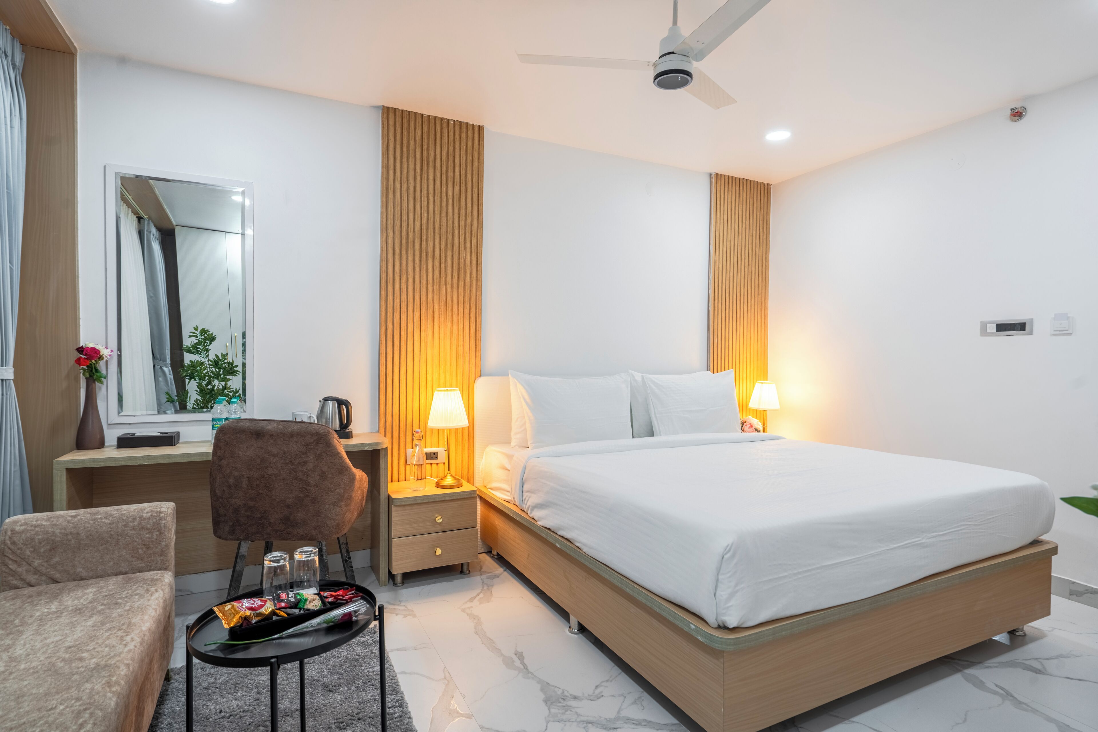 A modern hotel room with a large bed, bedside lamps, a desk with a chair, a small table with snacks, and a ceiling fan.