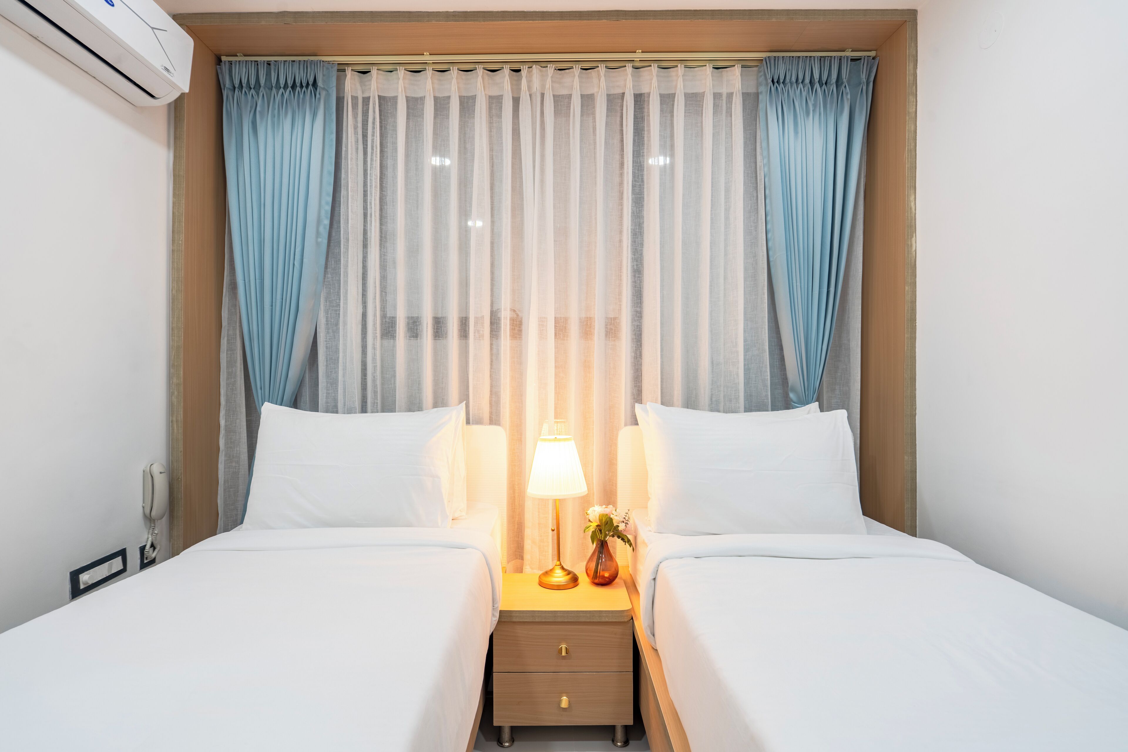 A hotel room with two beds, a bedside table with a lamp, and a small vase with flowers.