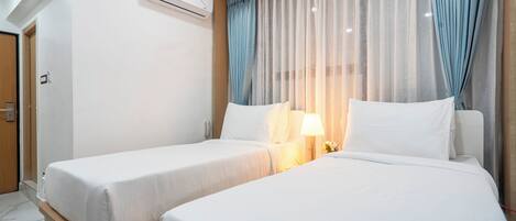 Una habitación de hotel con dos camas, una mesita de noche con lámpara y un ventanal con cortinas translúcidas.