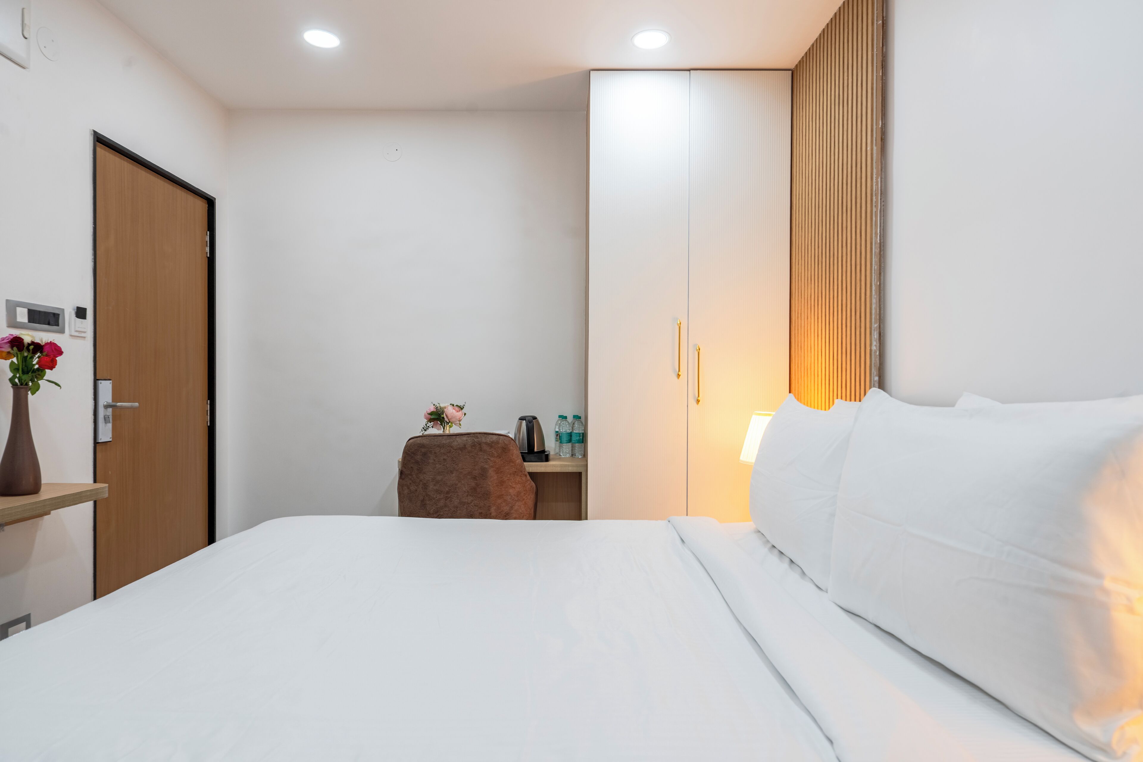 A hotel room with a bed, a desk with a chair, a wooden door, and a wall-mounted light fixture.
