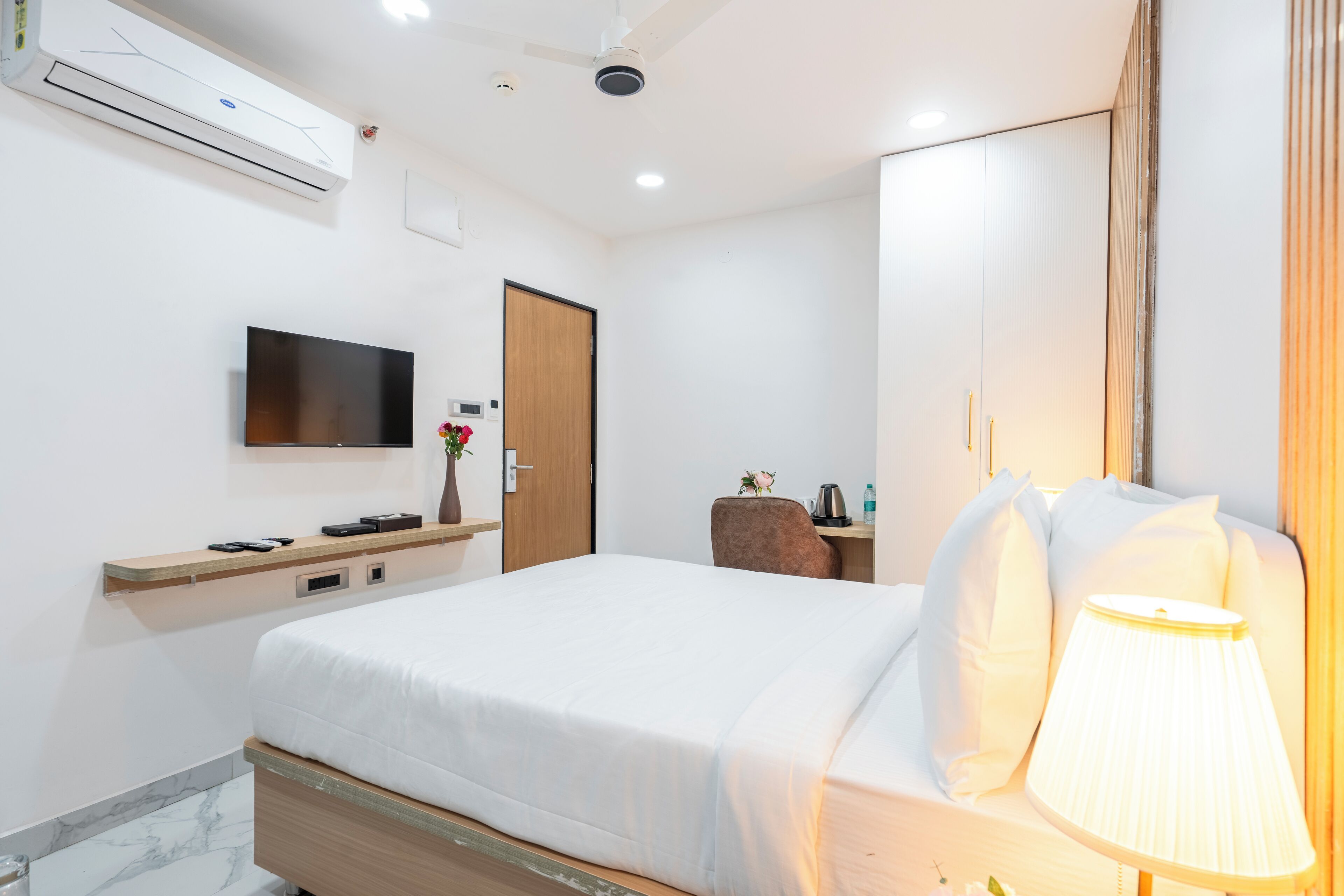 A hotel room with a bed, a TV mounted on the wall, a desk with a lamp, and a wardrobe.