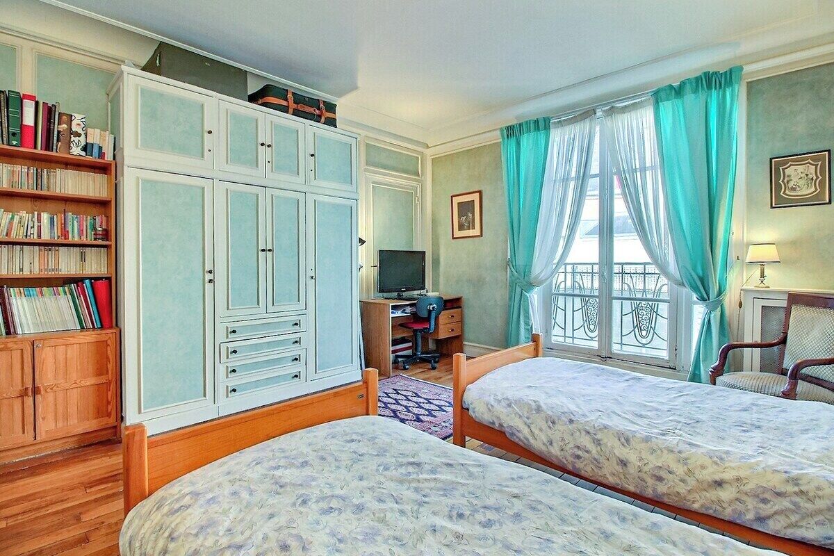 A bedroom with two beds, a desk with a computer, a bookshelf, and a window with curtains.