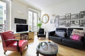 A living room with a black leather sofa, a red leather armchair, a glass coffee table, and a TV on a wooden cabinet. - Parisian Home | 2 bedrooms | Marais - Bastille (Paris)