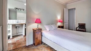A bedroom with a bed, a nightstand with a lamp, a wooden cabinet, and an open door leading to a bathroom. - Parisian Home | 2 chambres | Marais - Bastille (Paris)