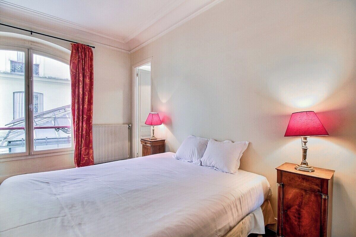 Habitación de hotel con cama, dos lámparas de noche, una ventana con vista a edificios y una cortina roja.