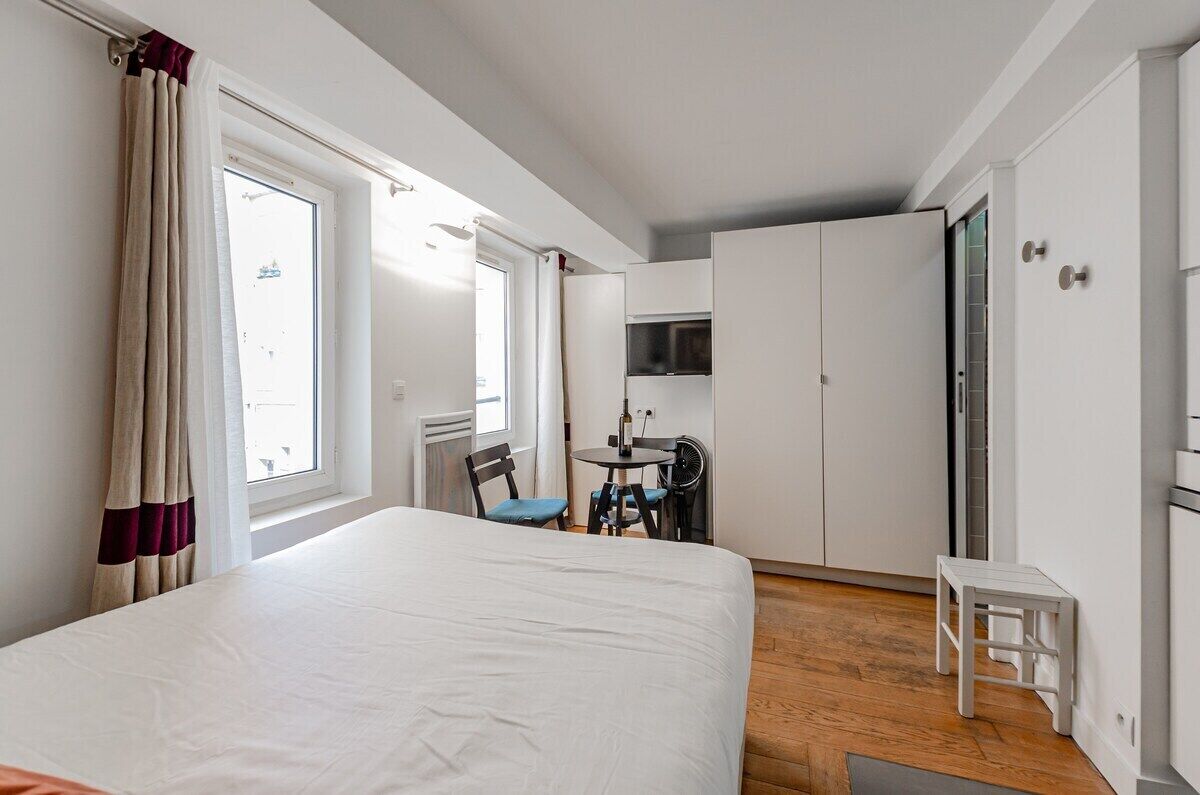 Hotellihuone, jossa on sänky, pieni pöytä tuoleineen, pieni keittiö sekä ikkuna, jossa on verhot.
