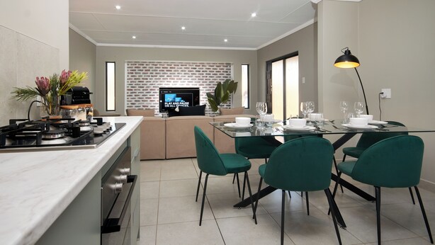 A modern kitchen with a built-in stove, a dining area with green chairs, and a television in the background.
