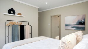 A bedroom with a bed, a hanging clothes rack, a shelf with decorative items, and a painting on the wall.
