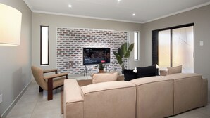 A modern living room with a sectional sofa, a flat-screen TV mounted on a brick-patterned wall, and a large window providing natural light.
