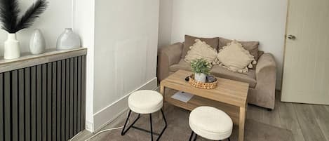 A small, cozy room with a wooden coffee table, two white stools, a beige sofa with decorative pillows, and a radiator.
