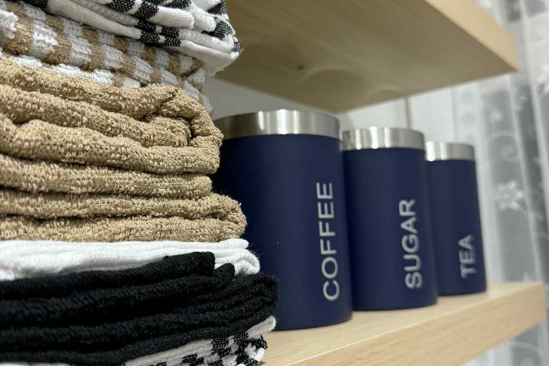A shelf with neatly stacked towels in various colors and three cylindrical containers labeled 'COFFEE,' 'SUGAR,' and 'TEA.'