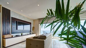 A modern living room with a large flat-screen TV, a beige sofa, and a large plant. - Mediterranean Luxury Stay with Sea Views & Jacuzzi (Xgħajra)