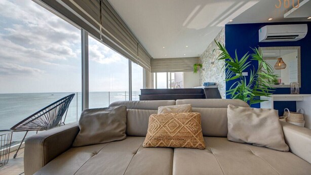 A modern living room with a large window overlooking the sea, a sofa with cushions, and a blue accent wall. - Mediterranean Luxury Stay with Sea Views & Jacuzzi (Xgħajra)
