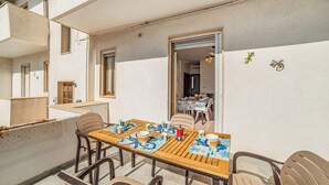 Uma área de refeições ao ar livre com uma mesa posta para quatro pessoas, rodeada por cadeiras, e vista para o edifício vizinho.
