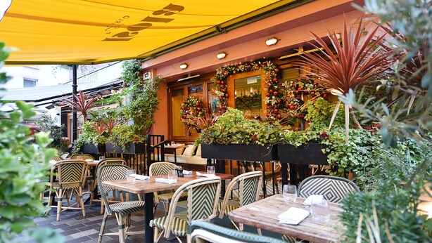 Un café al aire libre con mesas y sillas de madera, rodeado de macetas con plantas y un toldo amarillo.