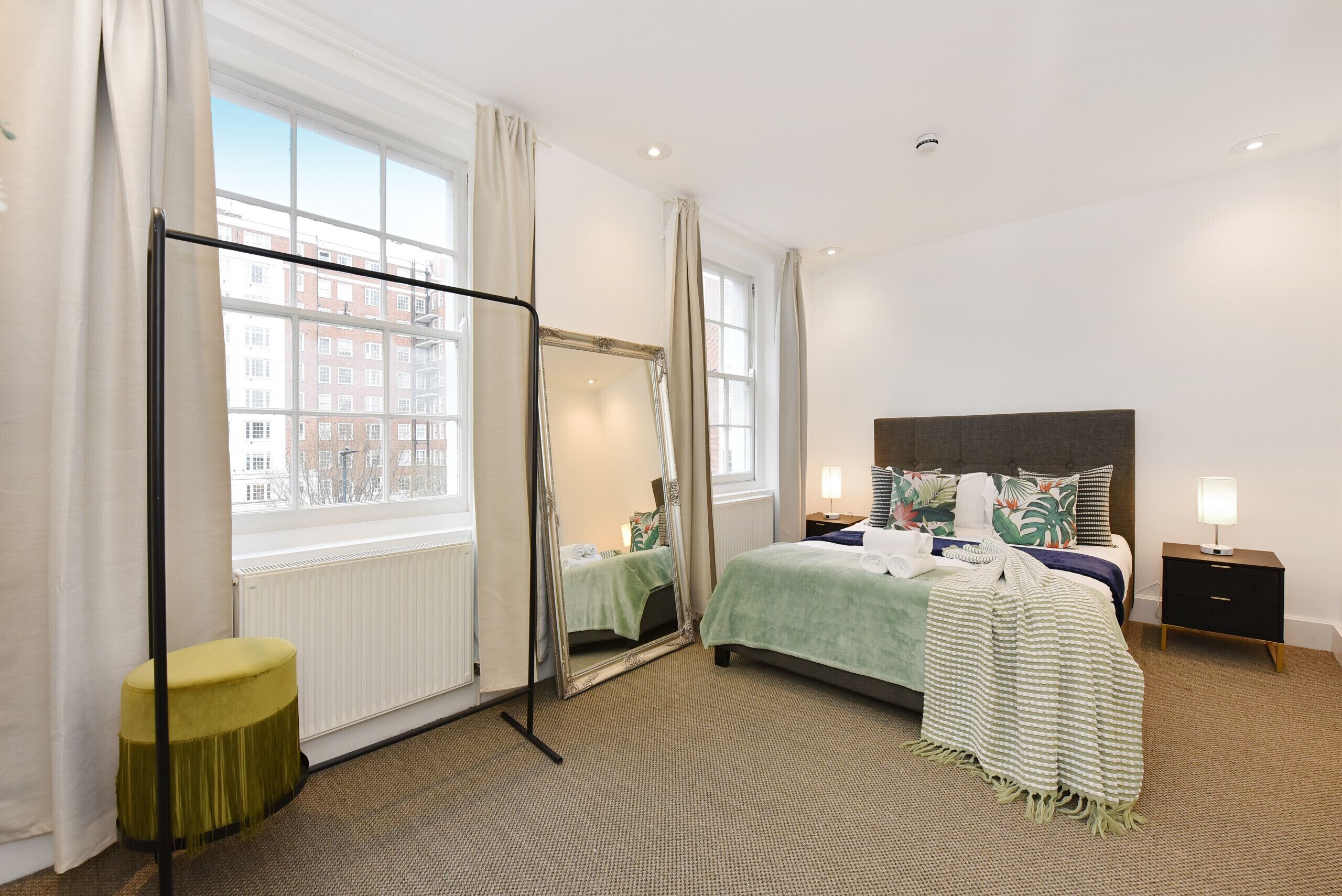 Ein Hotelzimmer mit einem großen Bett, einem Stehspiegel, einem kleinen Hocker, einem Nachttisch und Blick auf Gebäude durch das Fenster.