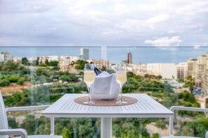 Una mesa con dos copas de vino blanco y una botella en un balcón con vistas a un paisaje urbano.