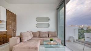 Una sala de estar moderna con un sofá seccional color beige, una mesa de centro de vidrio con una planta y una vista de la ciudad a través de amplias ventanas.