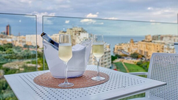 Una mesa con una botella y dos copas de vino blanco en un balcón con vistas a un paisaje urbano.