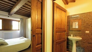 Una habitación con cama, puertas de madera, baño con lavamanos y espejo, y pared de ladrillo.