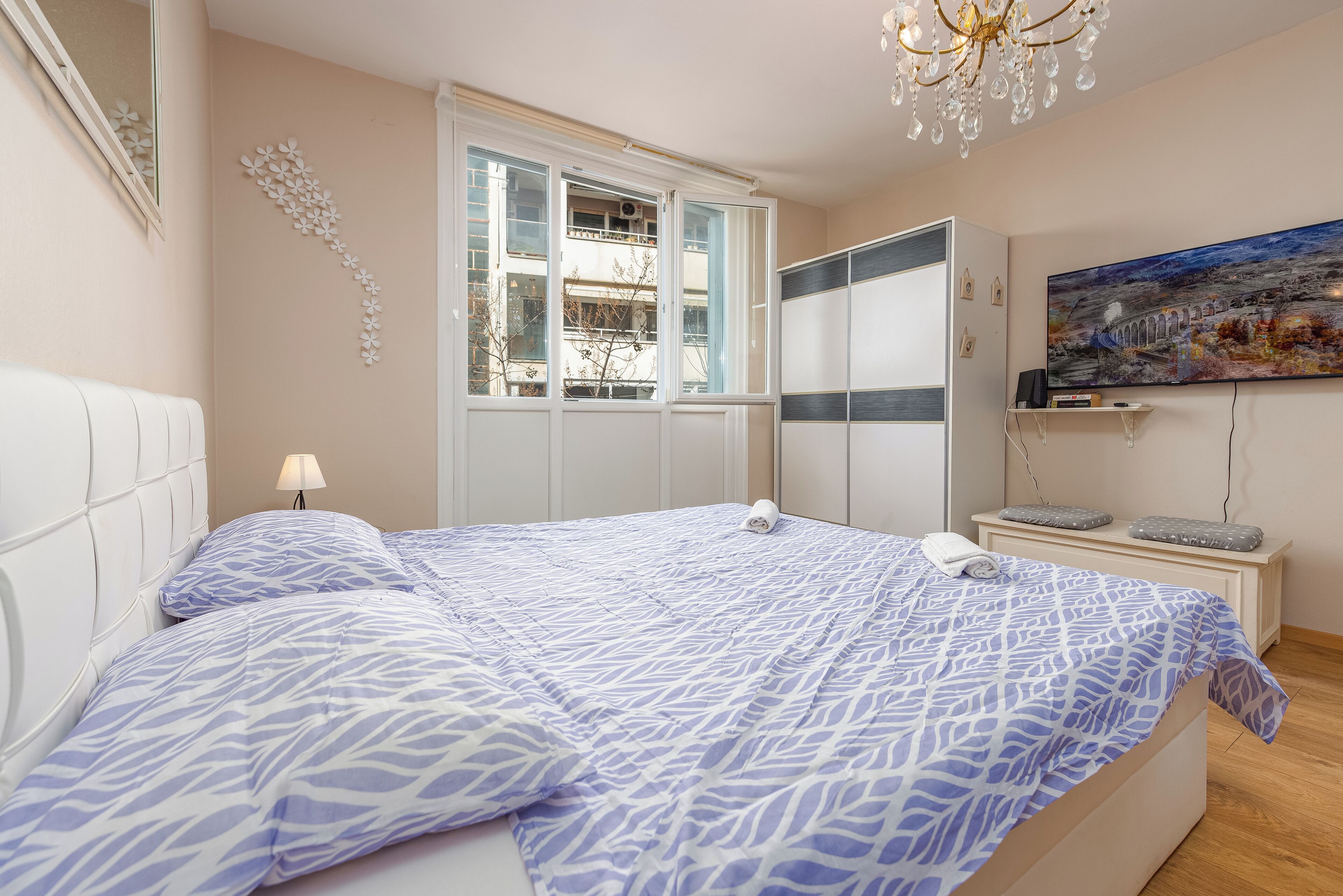 A bedroom with a bed, a window, a bedside table, a TV, and a painting on the wall.