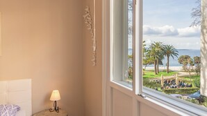 A room with a bed, a lamp, and a window overlooking a beach with palm trees and people. - Apartment Oasis with Sea View (Zadar)