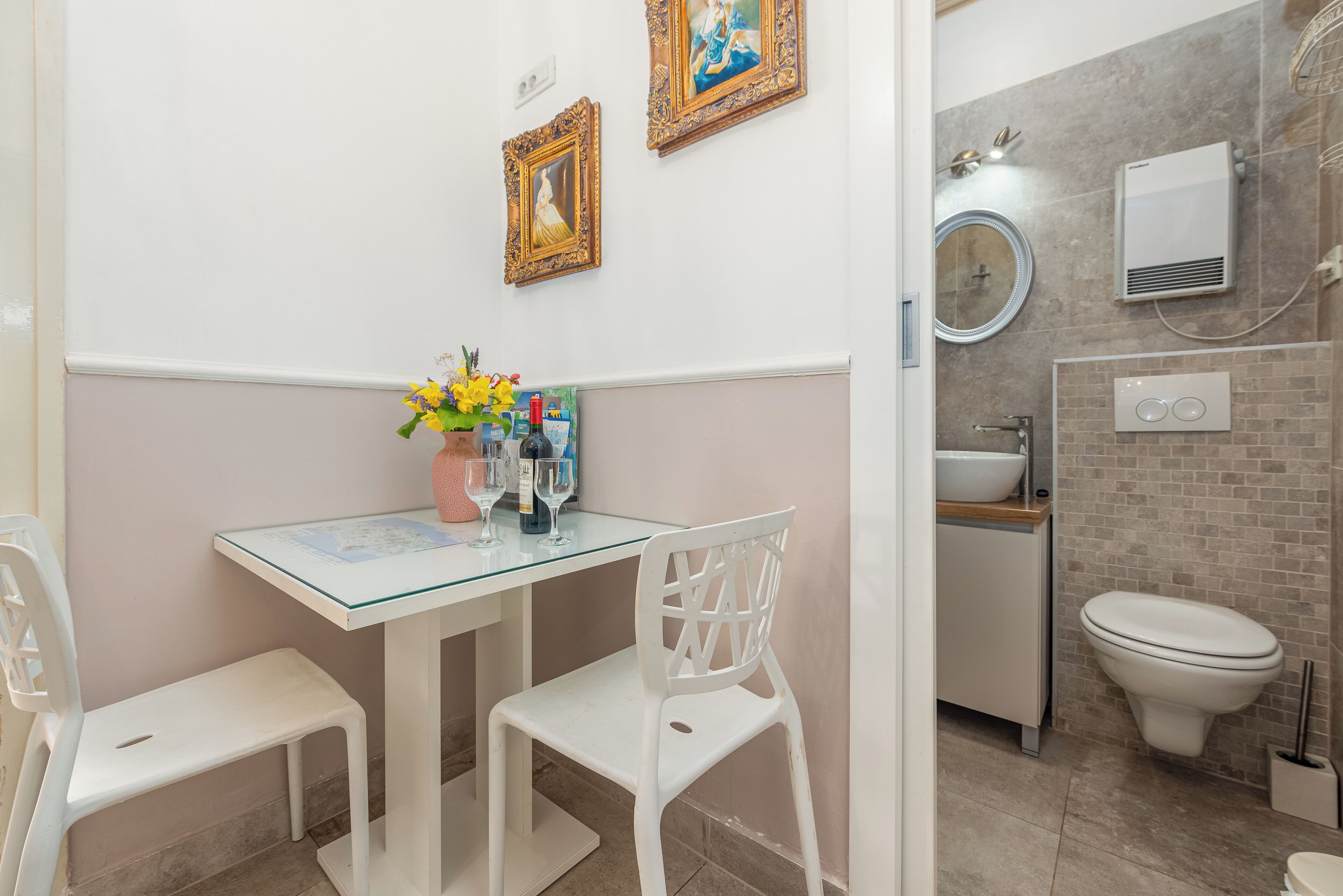 Un baño con lavamanos, inodoro y tocador blancos. Hay una pequeña mesa con un florero y un espejo encima.