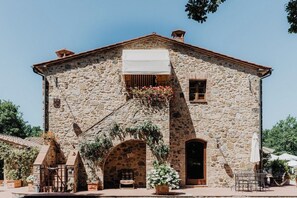Un edificio de piedra con un arco central, un pequeño balcón con toldo blanco y mesas al aire libre.