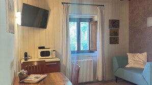 A cozy room with a wooden dining table, chairs, a sofa, a TV mounted on the wall, and a window with curtains. - Standard two-room flat Nr 3 (Radicondoli)