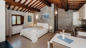 Quarto com teto de madeira, uma cama, janela com cortinas, um quadro na parede e uma área de cozinha com fogão.