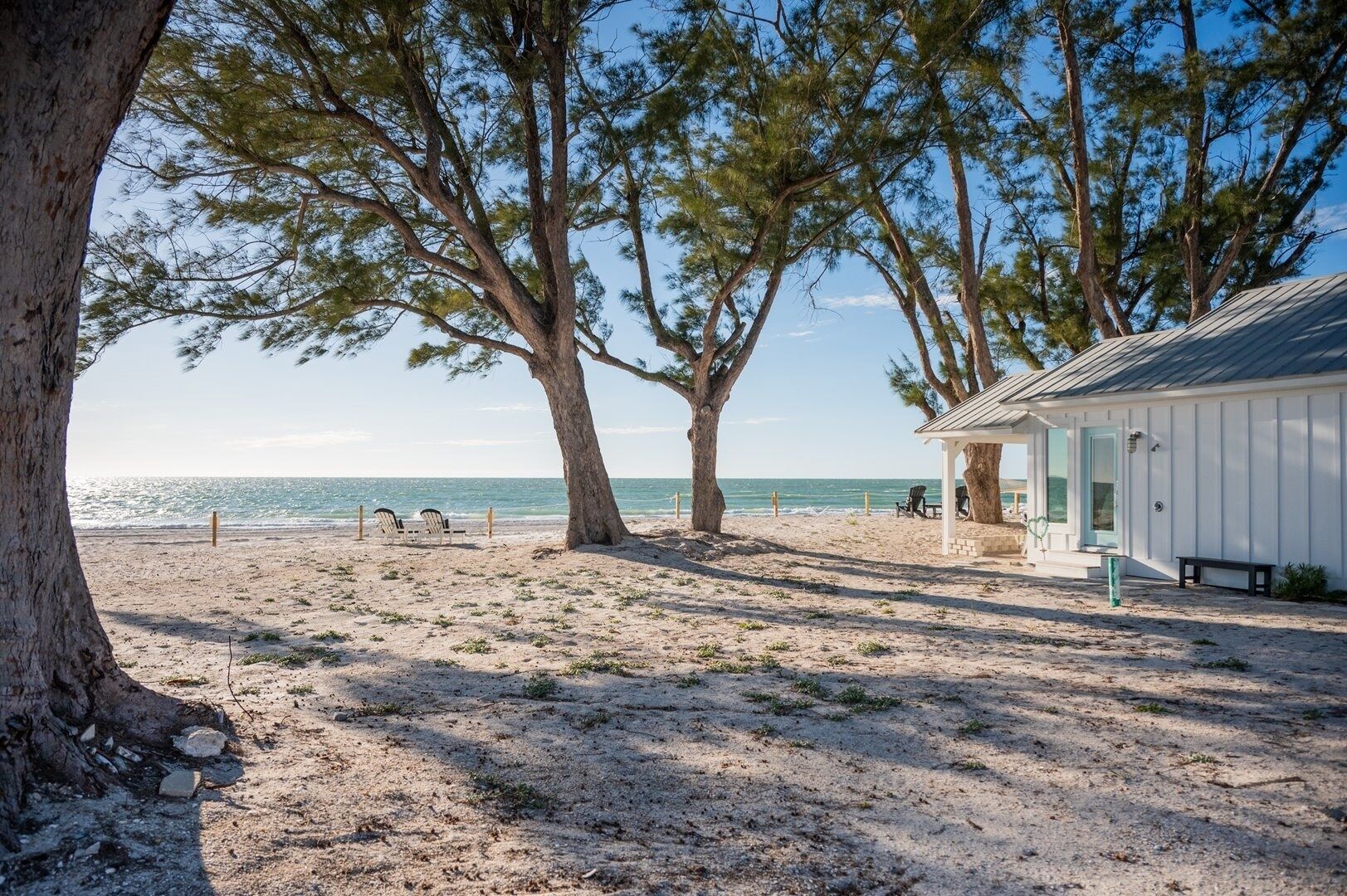 A beach scene with a small white building, two chairs, and a bench near the water.