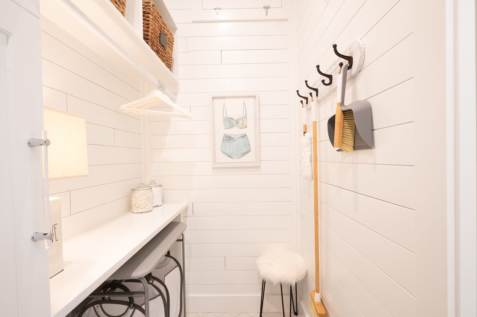 Un baño estrecho con paredes de madera blanca, un lavabo blanco con espejo, una obra de arte enmarcada y una escoba apoyada en la pared.