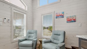 A room with two light grey armchairs, wooden flooring, white paneled walls, and two windows with blinds. - Sailfish Rv Resort 2 Home (Port Aransas)