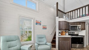 A spacious living area with two light blue armchairs, a wooden floor, a kitchen with modern appliances, and a staircase leading to an upper level. - Sailfish Rv Resort 2 Home (Port Aransas)